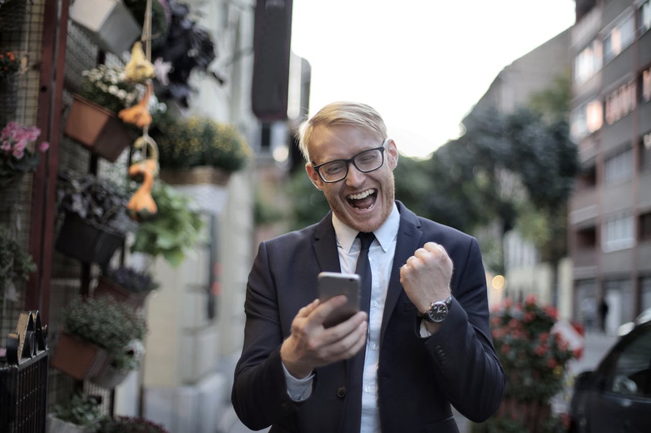 Excited young man in formal wear and eyeglasses clenching fist and rejoicing victory while using mobile phone in modern city street