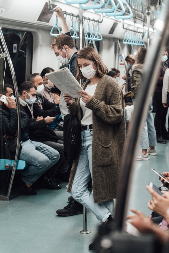 Anonymous woman in sterile mask reading newspaper near crop passengers on subway during COVID 19 pandemic